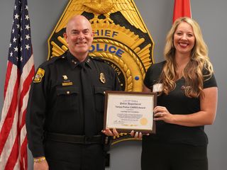 Man in uniform next to woman in dress holding a certificate in front of flags and large law enforcement shield on the wall.
