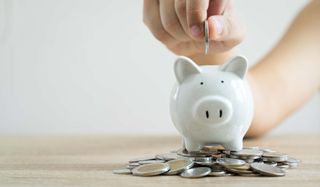 hand dropping coins into a small white piggy bank. More coins around bank on table.