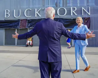 A man in a navy suit with his back to the camera, arms open wide as a woman in light blue jacket and pants walks towards him. On the building is the word "Buckhorn" with the man's head covering the H.