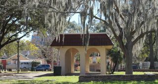 A tan pavilion with two arched openings on each side situated in park with large oaks overhanging.