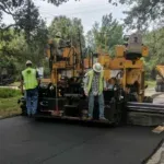 Heavy machinery used for resurfacing roads with a crew working in a neighborhood. Large oaks can be seen hanging over the road and in background.