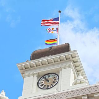 US flag, city flag, Pride flag flying above Tampa City Hall on a clear blue day with small tufts of white clouds.