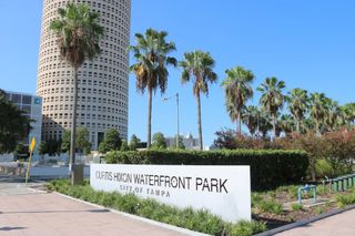 Sign for the entrance to Curtis Hixon Park with tall round building in background and a line of palm trees.