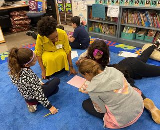 A Black woman in a yellow dress on a blue carpet in a classroom watching students working in a group on the floor.