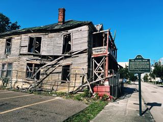 A crumbling two story wood structure with a historical marker on the sidewalk out front.