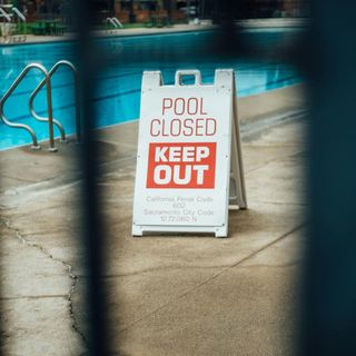 A view through bars at a sign that reads "Pool Closed. Keep Out." Portion of pool can be seen behind the sign.