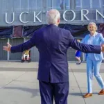 A man in a navy suit with his back to the camera, arms open wide as a woman in light blue jacket and pants walks towards him. On the building is the word