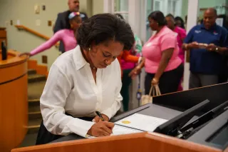 A young Black woman in a white blouse signs documents at a podium. A small crowd out of focus is behind her.