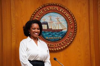 A young Black woman in white shirt stands in front of the City of Tampa seal that hangs behind council dais.