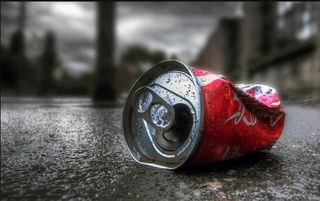 A dented Coca Cola can on the road with a city scape out of focus in the background.