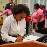 A young Black woman in a white blouse signs documents at a podium. A small crowd out of focus is behind her.